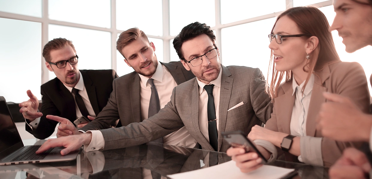 Een groep van vijf professionals in zakelijke kleding voert een levendige discussie aan een glazen vergadertafel met een laptop, waarbij ze samenwerking en concentratie uitstralen.