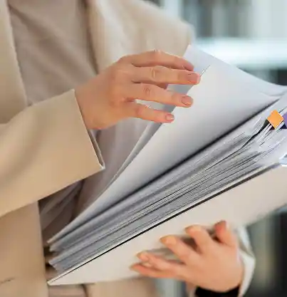 Een persoon in een beige pak bladert door een grote stapel documenten met gekleurde bladwijzers, wat een indruk van concentratie en organisatie in de kantooromgeving wekt.